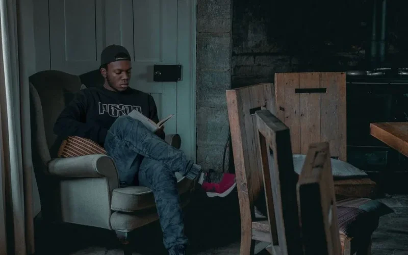 A man relaxes indoors, reading a book in a cozy chair with natural light.