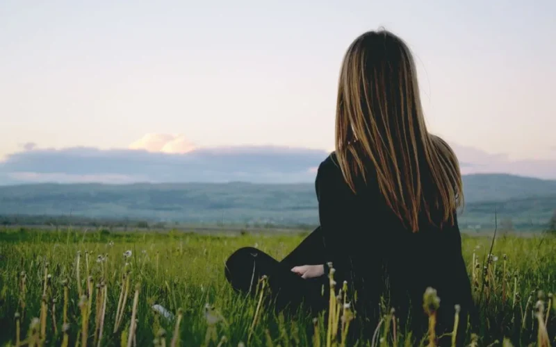Woman enjoying peace in a lush green field with a scenic mountain view during sunset.