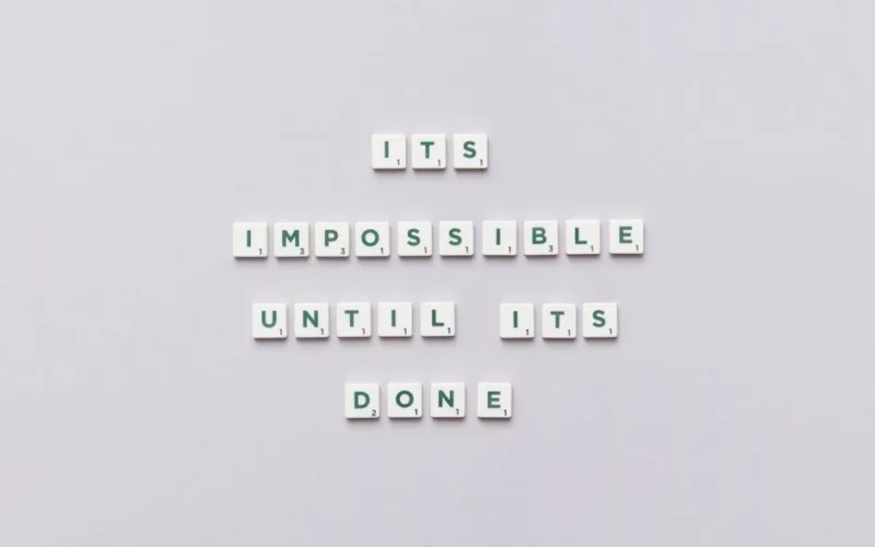 Motivational phrase 'It's impossible until it's done' arranged with Scrabble tiles on a gray background.