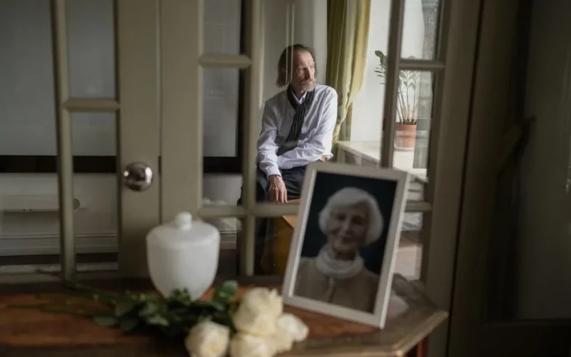 A senior man sits reflectively indoors with a photo and flowers, evoking a sense of mourning.