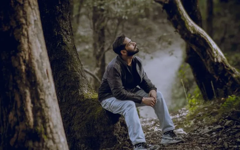 A man sitting thoughtfully under a tree in a tranquil forest environment.