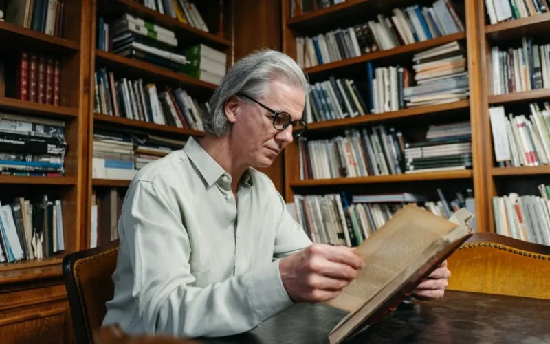An elderly man with gray hair reading a book in a library surrounded by bookshelves.