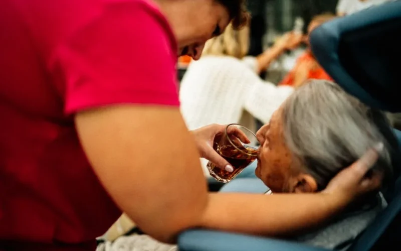 A caregiver helps an elderly woman drink tea, showcasing empathy and support.