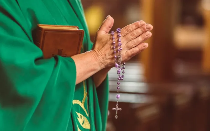 A priest dressed in green robes holds a rosary and Bible, symbolizing faith and prayer.