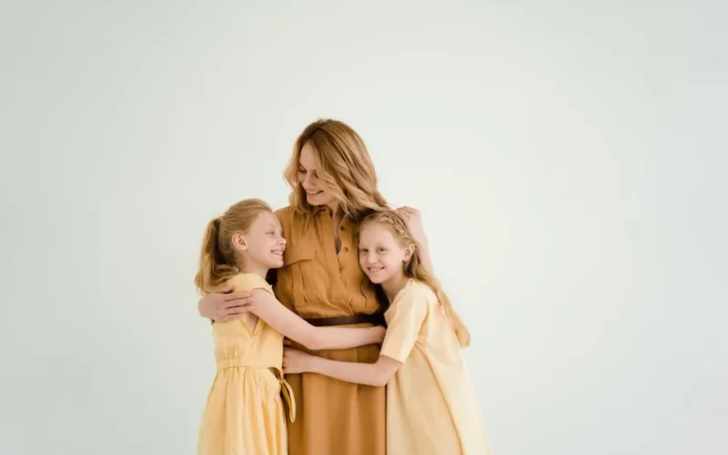 A mother lovingly embraces her two daughters against a soft, light background, symbolizing family love and togetherness.