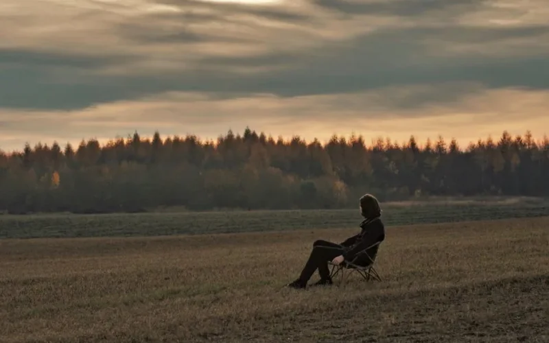 A solitary person sits on a chair in a tranquil countryside field at sunset, evoking peace.