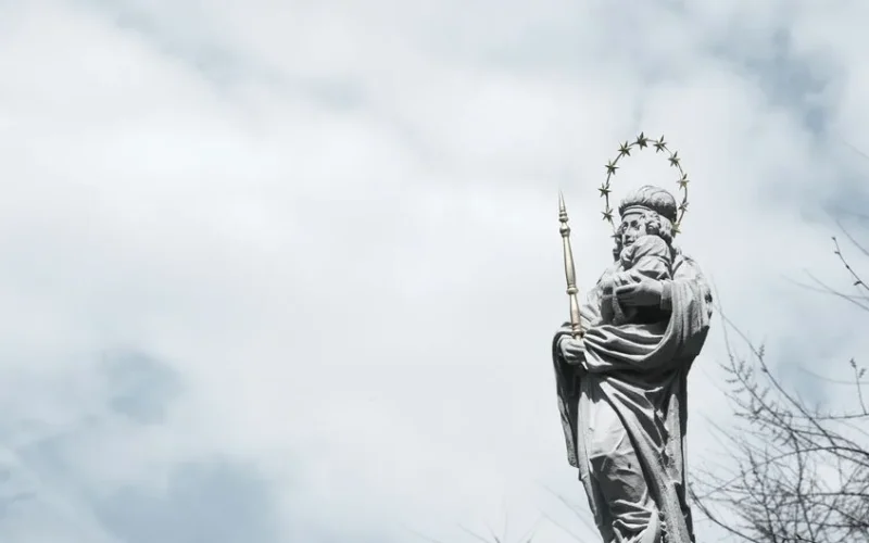 A striking statue of a saint with a halo and scepter set against a dramatic cloudy sky.
