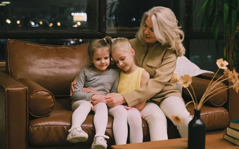 Grandmother lovingly embraces two granddaughters on a cozy leather sofa indoors.
