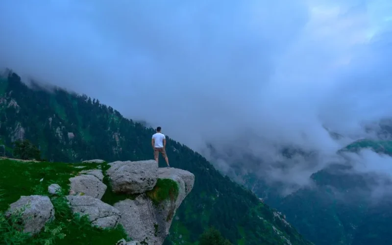 A man stands on a cliff, surrounded by misty mountains and lush greenery.
