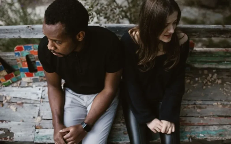 A couple sitting apart on a park bench, expressing emotions. Outdoors setting.