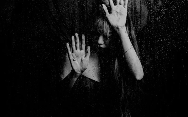 Dramatic black and white portrait of a woman behind a rainy window, evoking a moody atmosphere.
