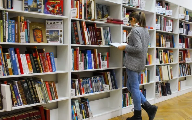 A young woman stands reading a book in a well-stocked library.