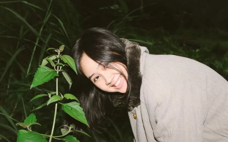 A woman with a joyful smile in a cozy coat is outdoors at night near lush green plants.
