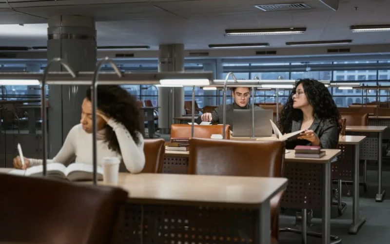 College students studying in a modern library setting, concentrating on books and laptops.