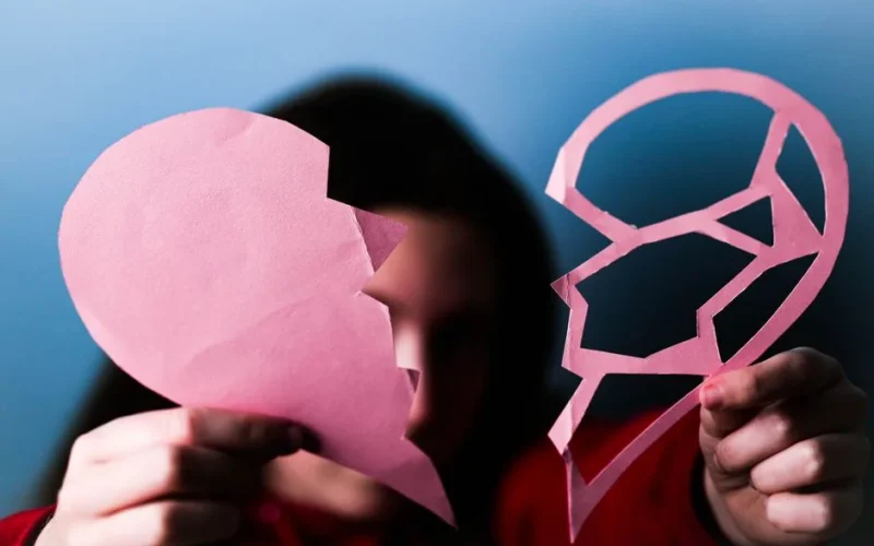 Close-up of hands holding a pink broken paper heart, symbolizing love and heartbreak.