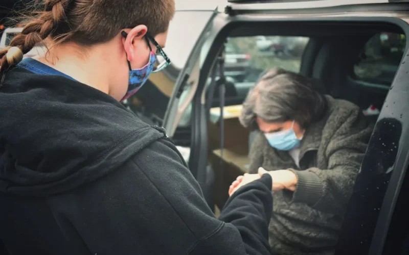 A person assists an elderly man wearing a mask by their car, symbolizing care during the pandemic.