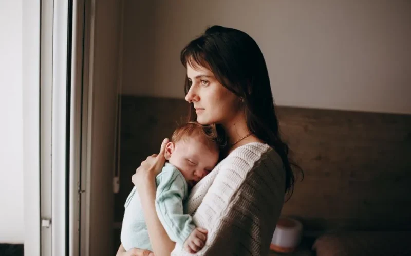 A serene moment of a mother holding her sleeping baby near a window, indoors.
