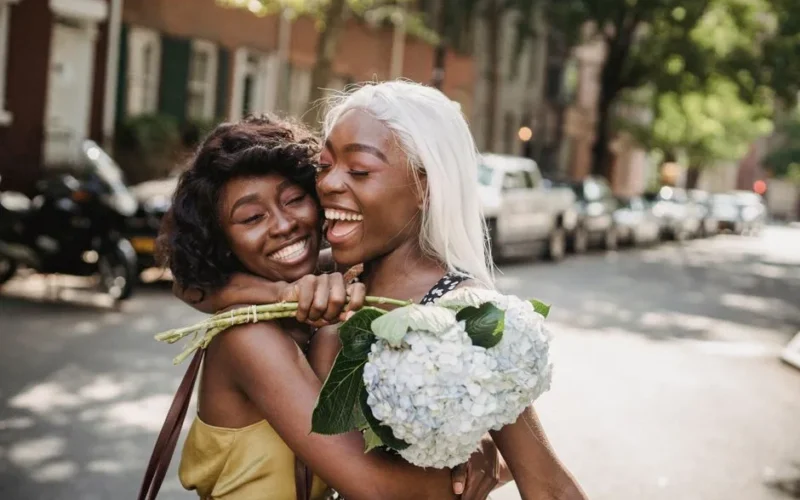 Two smiling women embrace on a sunny urban street, sharing joyful moments with a bouquet.