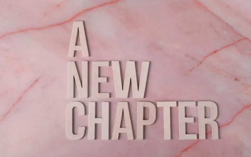 Wooden letters spell 'A New Chapter' on a pink marble background, symbolizing beginning.