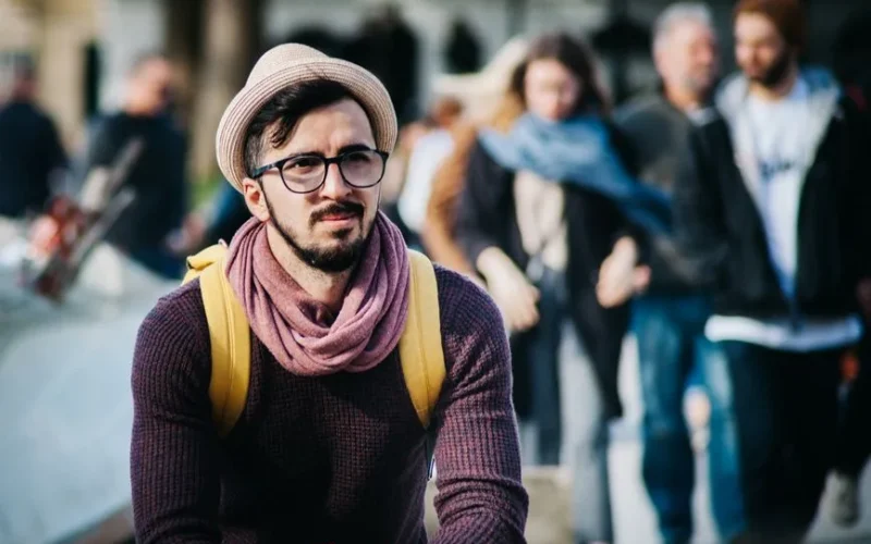 A stylish man in a trendy outfit stands out on a busy city street with various people in the background.