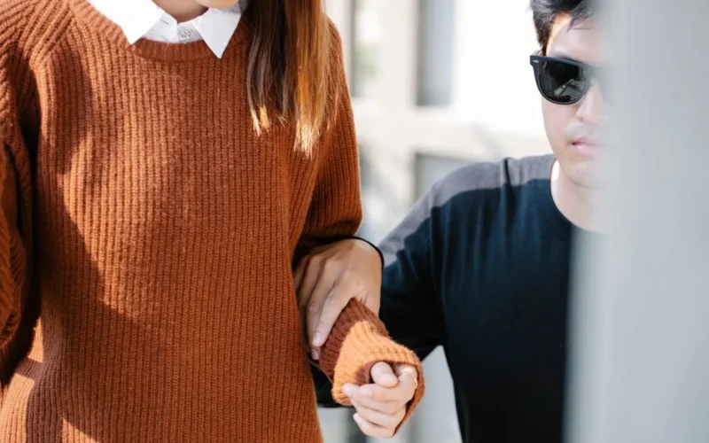 A man wearing sunglasses guides a visually impaired woman outdoors, highlighting care and assistance.