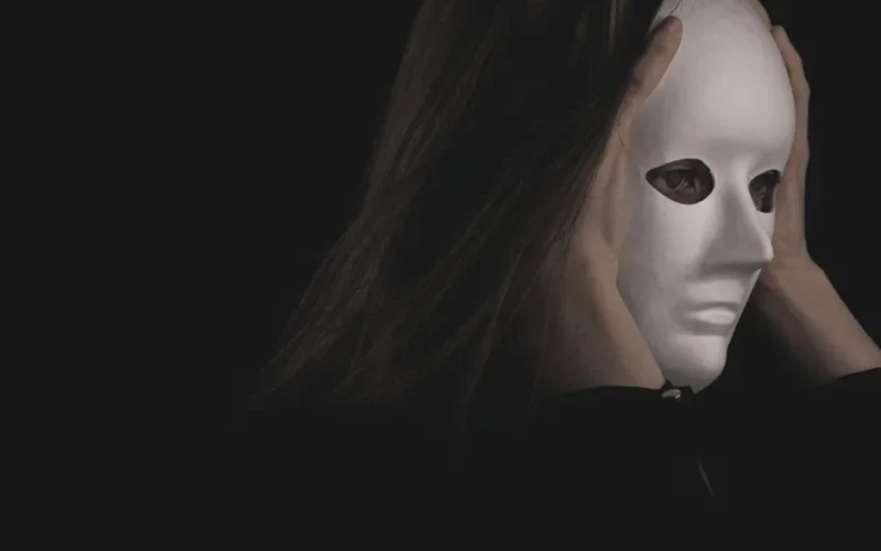 A person wearing a white mask held with both hands against a dark background, creating a mysterious atmosphere.