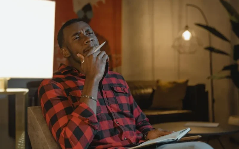 A thoughtful man in a plaid shirt contemplating indoors with a notebook.
