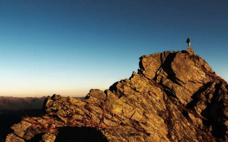 A lone hiker stands triumphantly atop a rocky mountain peak at sunrise, embracing nature and the view.