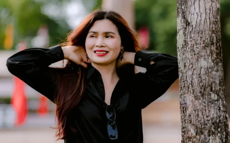 A woman smiling and relaxing outdoors, leaning against a tree in a sunny park setting.