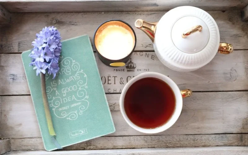A cozy tea setup featuring a cup, teapot, notebook, and candle on a wooden tray.