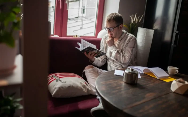 A man sits in a cozy room, reading a book by the window with a thoughtful expression.