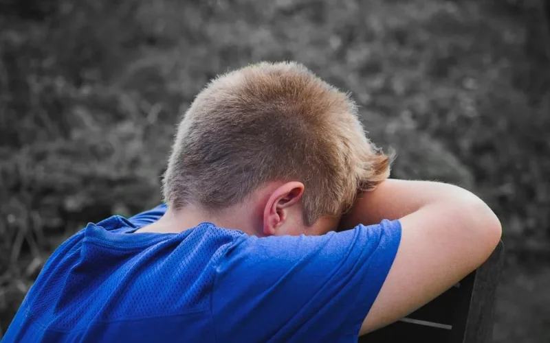 A young boy in a blue shirt rests his head on his arms outdoors, appearing sad and contemplative.