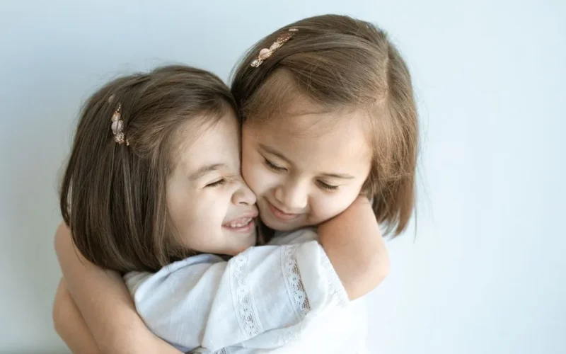 Two young girls embracing warmly, displaying sibling love and happiness against a plain background.