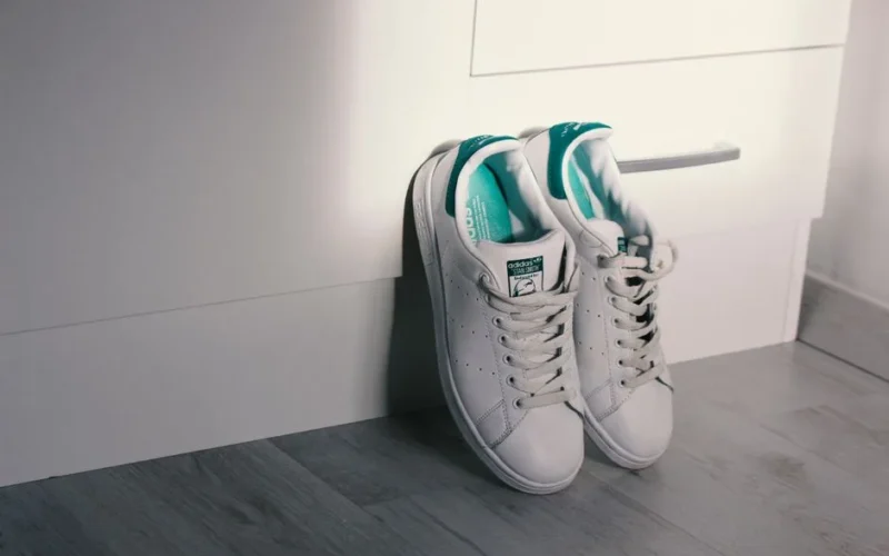 White Adidas sneakers with teal accents leaning against a drawer on a wooden floor.