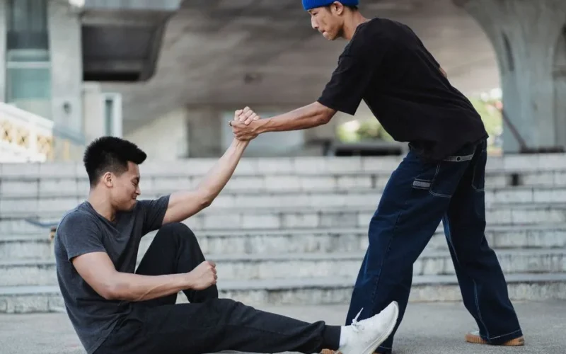 Side view Asian man in street wear holding friends hand and helping to get up from ground on sunny street
