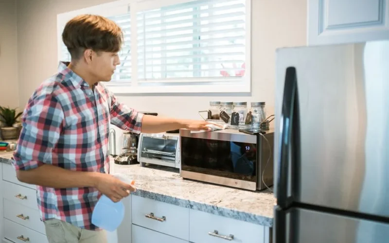 A man is cleaning a microwave in his modern kitchen using a spray bottle and cloth.