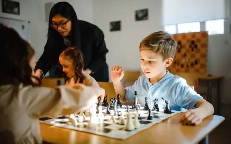 Young children playing chess under adult supervision in a classroom setting.