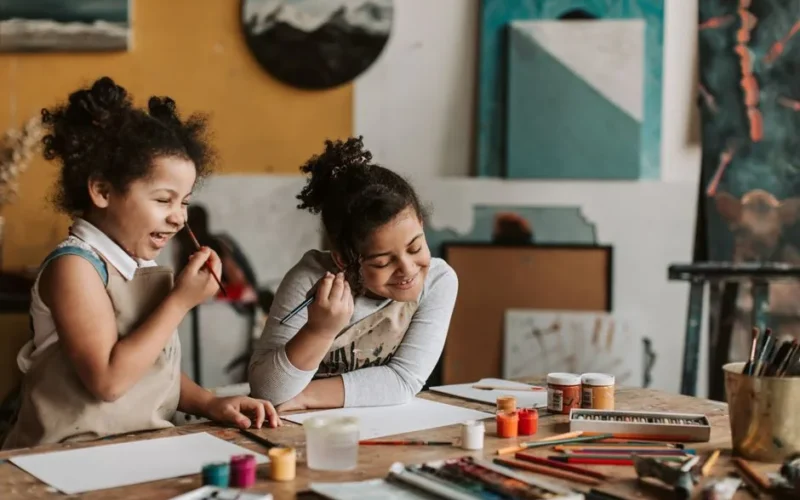 Two young girls happily painting during an indoor art workshop, expressing creativity and learning.