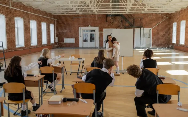 Teacher comforts a student in a classroom as other students watch. Captures school tension and support.