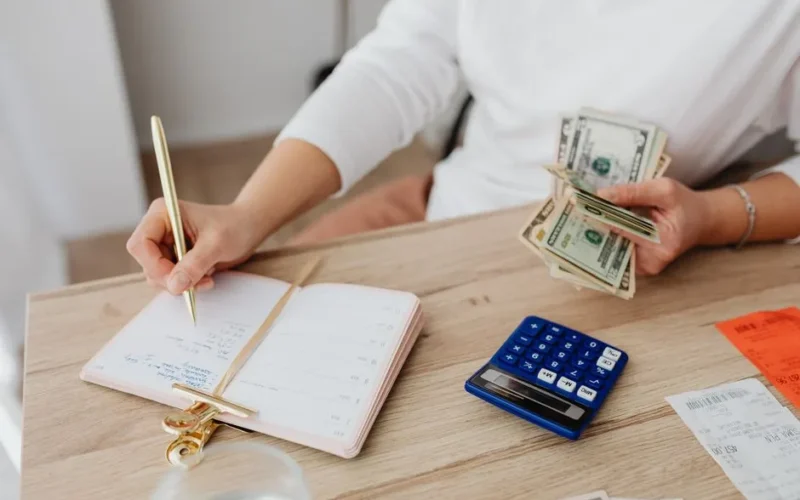 Adult holding cash and writing in planner while using a calculator at home.