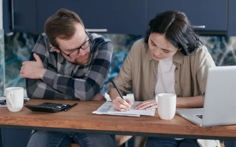 Adult couple working together on documents at a desk, with laptop and calculator nearby, in a home setting.