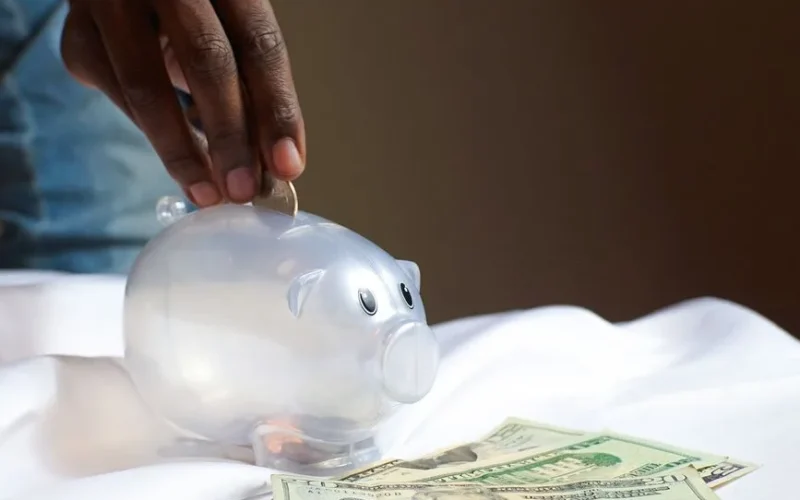 A hand placing a coin in a piggy bank with dollar bills nearby signifying savings and finance.