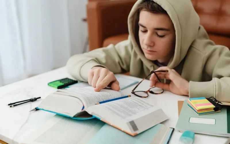 A teenager in a hoodie studying with books and notes on a table, focused on learning indoors.
