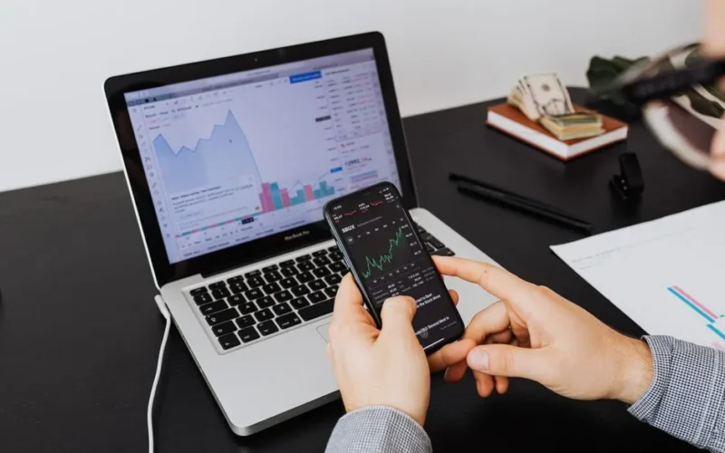 Person analyzing stock market data on a laptop and smartphone indoors.