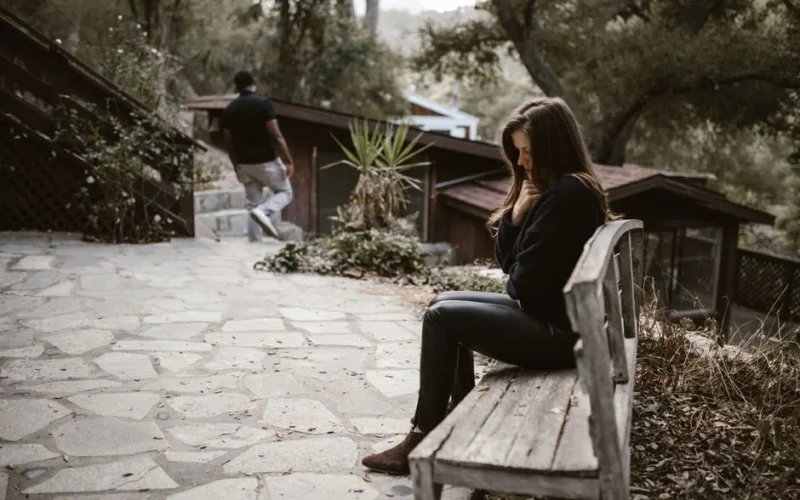 A woman sits pensively on a bench outside after a breakup, while a man walks away.