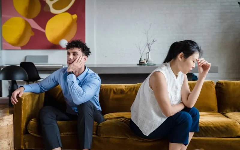 A couple sits apart on a sofa, looking unhappy and deep in thought, reflecting relationship conflict.