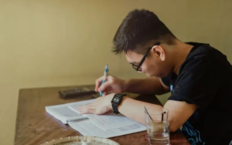 A young man concentrates on studying at his desk, taking notes indoors.