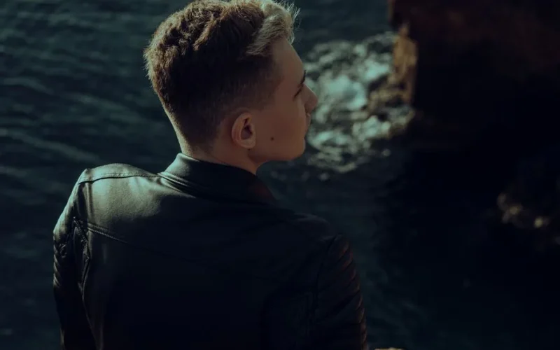 Young man in leather jacket looking thoughtfully over the sea from a rocky edge.