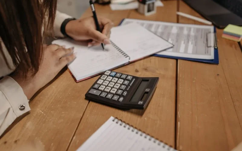 Woman writing notes with a calculator on a wooden desk, focusing on work and accounting.