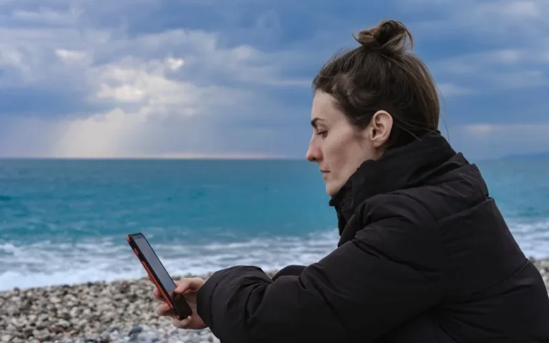 A woman in a black jacket sitting on a pebble beach, using a smartphone with an ocean backdrop.